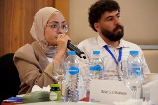 A conference attendee wearing a beige hijab and glasses speaks while another attendee with a beard listens. They are seated at a table.