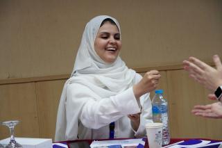 A woman in a white hijab and blouse smiles warmly as she engages in conversation during a conference. She gestures towards another person whose hand is partially visible.