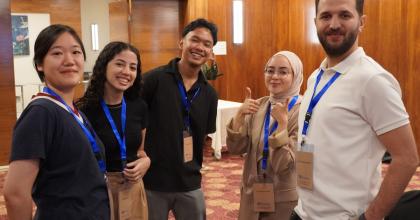 A group of five people wearing conference badges and lanyards stand together in a room with wood-paneled walls, smiling and posing for the camera.